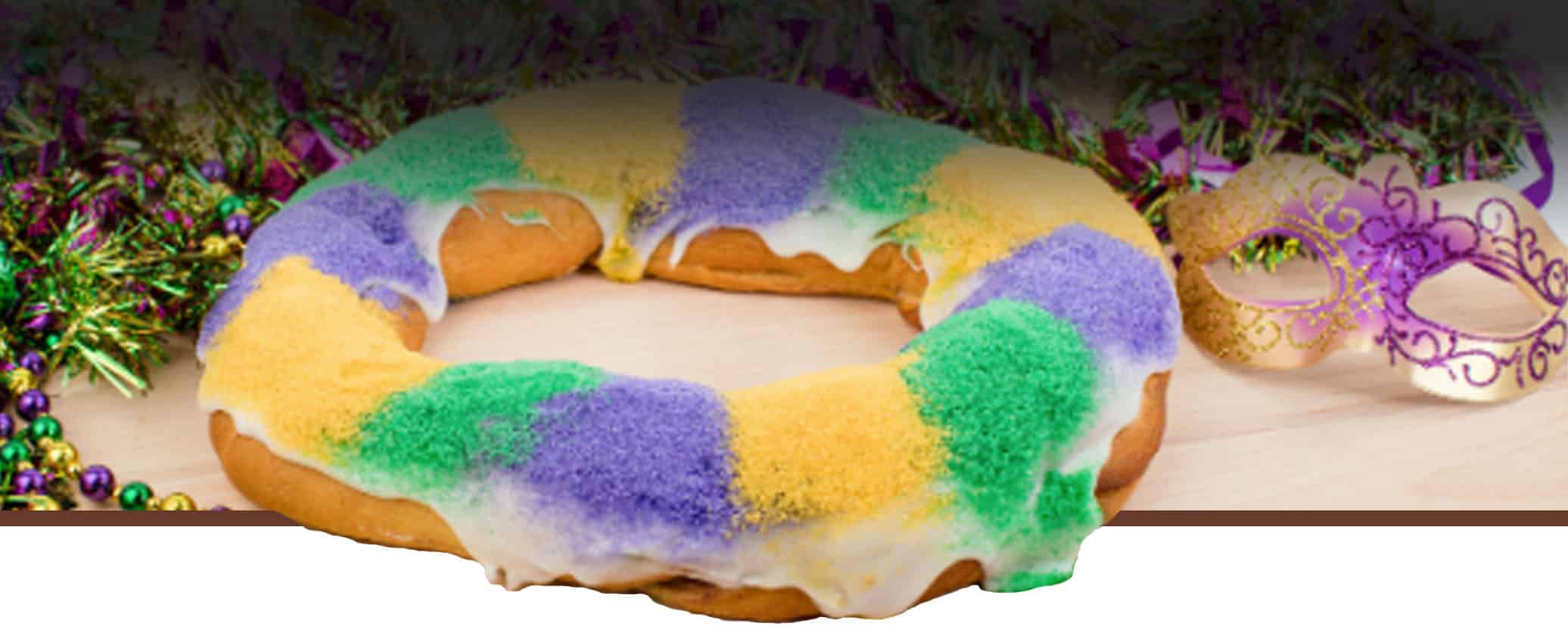 A King Cake with green, purple, and yellow sugar, partially glazed, sits on a table next to colorful beads and a decorative masquerade mask.